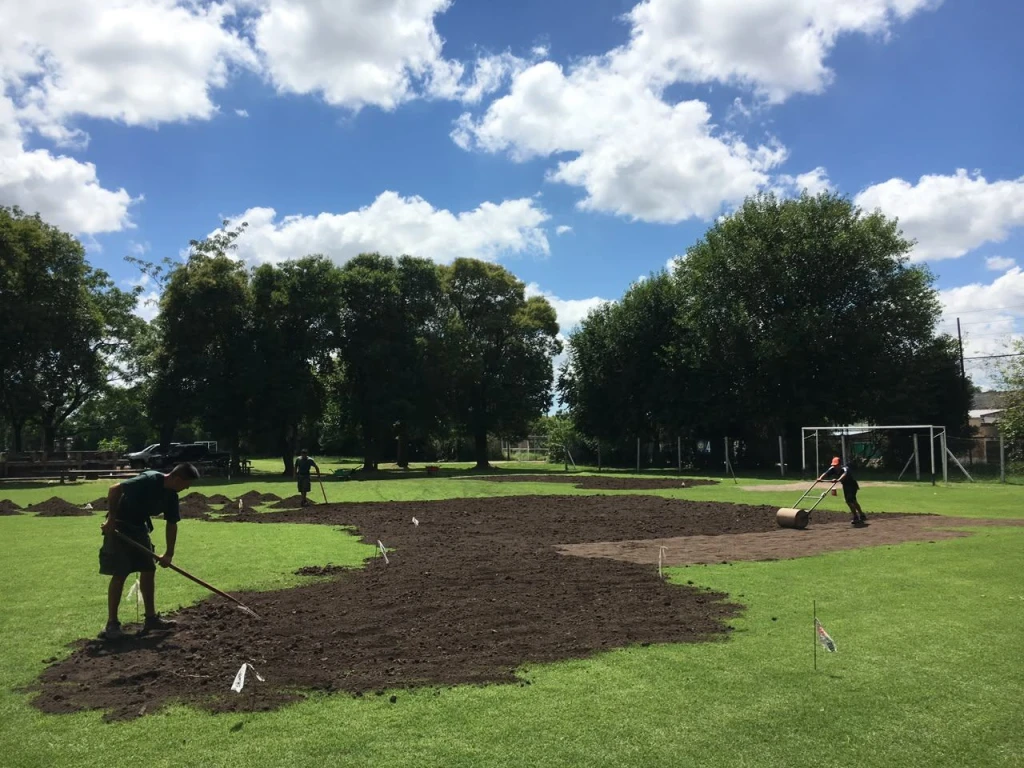 Cuadrilla de Paisar desparramando tierra con rastrillo y compactando con rodillo en la cancha del Instituto Inmaculada, Castelar