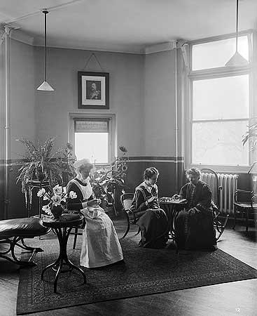 A nurse with two female patients playing draughts on The King Edward VII ward at The Royal London Homeopathic Hospital. A portrait of King Edward is hanging above a window behind the three women.